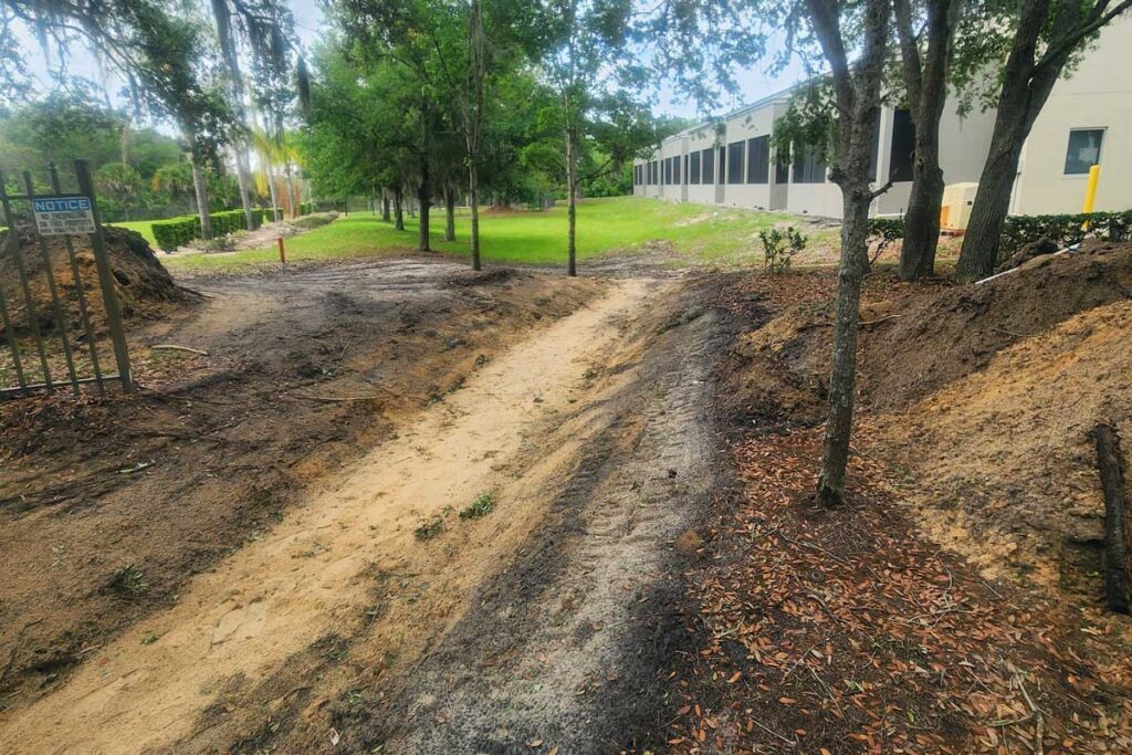Dirt pathway under construction beside a building, with trees and grass in the background and a "NOTICE" sign on a gate to the left.