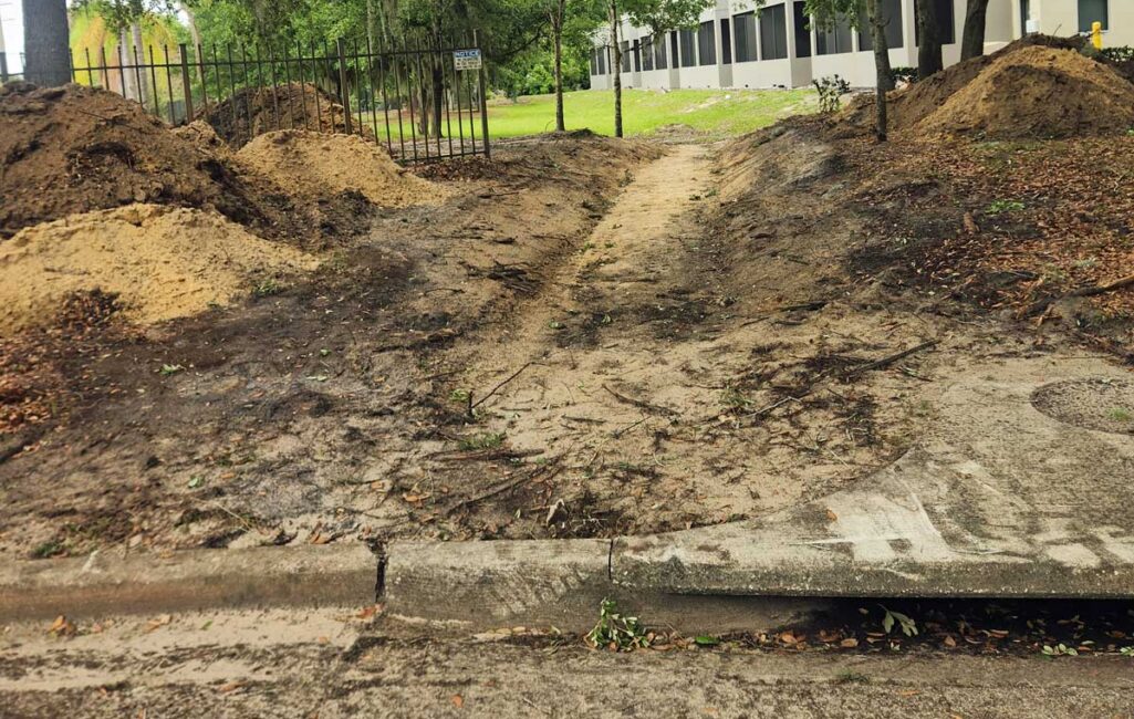 A dirt path worn into the ground leads from a sidewalk through a gap in a fence, with piles of soil on both sides and a building visible in the background.