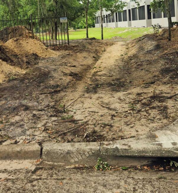A dirt path worn into the ground leads from a sidewalk through a gap in a fence, with piles of soil on both sides and a building visible in the background.