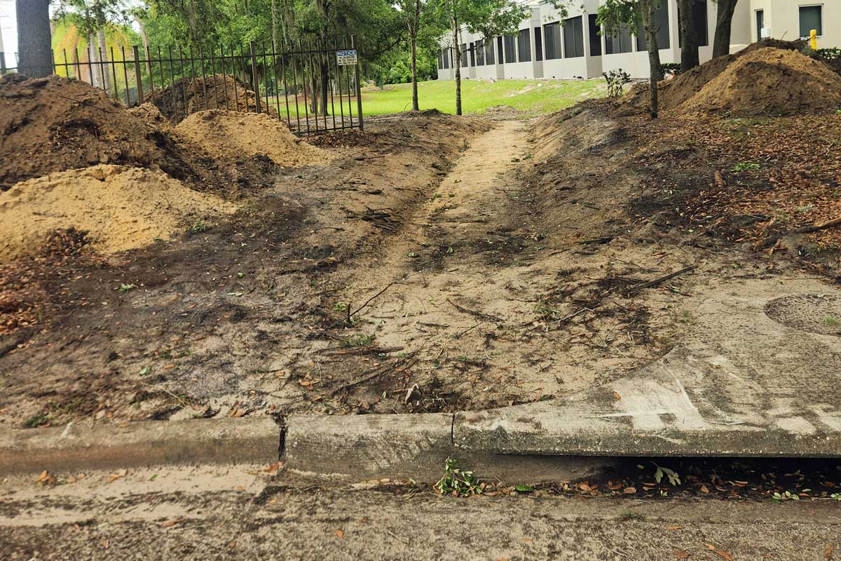 A dirt path worn into the ground leads from a sidewalk through a gap in a fence, with piles of soil on both sides and a building visible in the background.