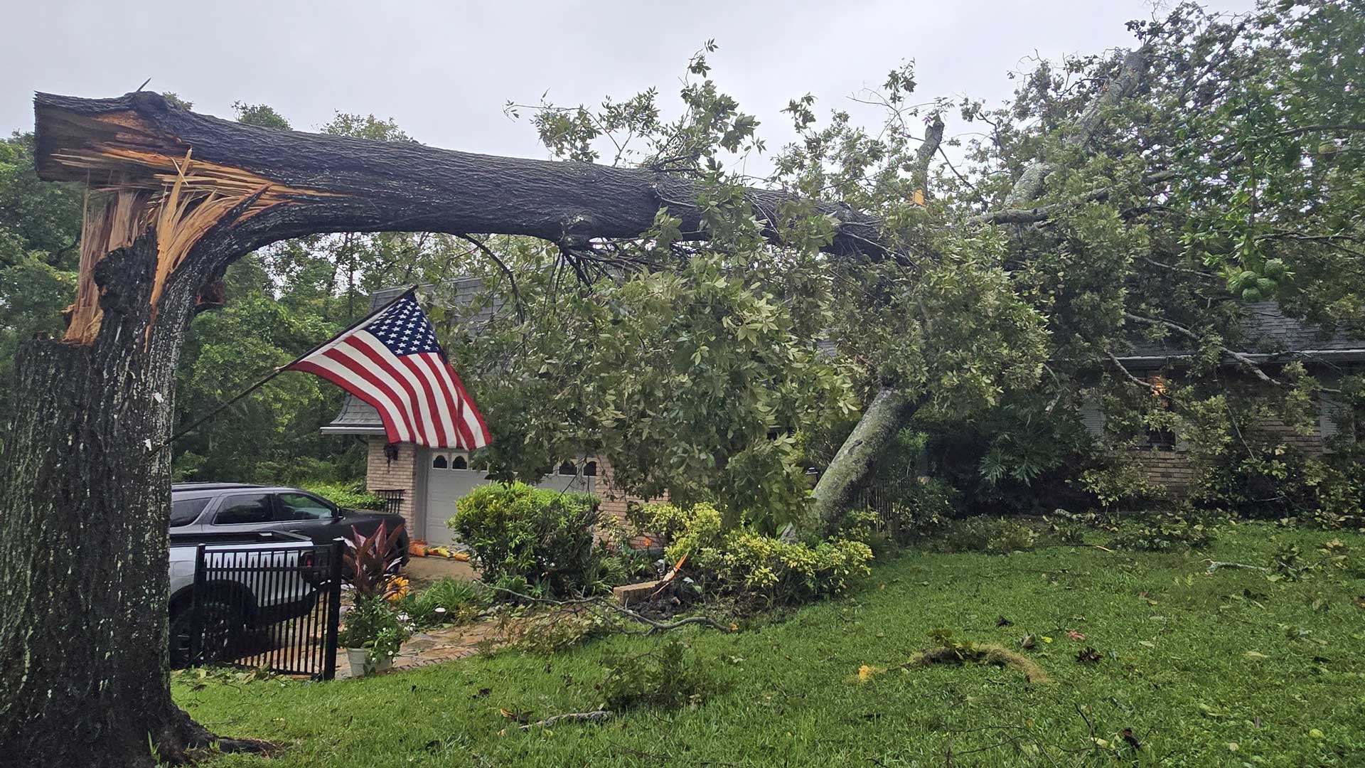 A large tree has fallen and is split in half, resting on a house and car in a residential yard. An American flag hangs nearby.