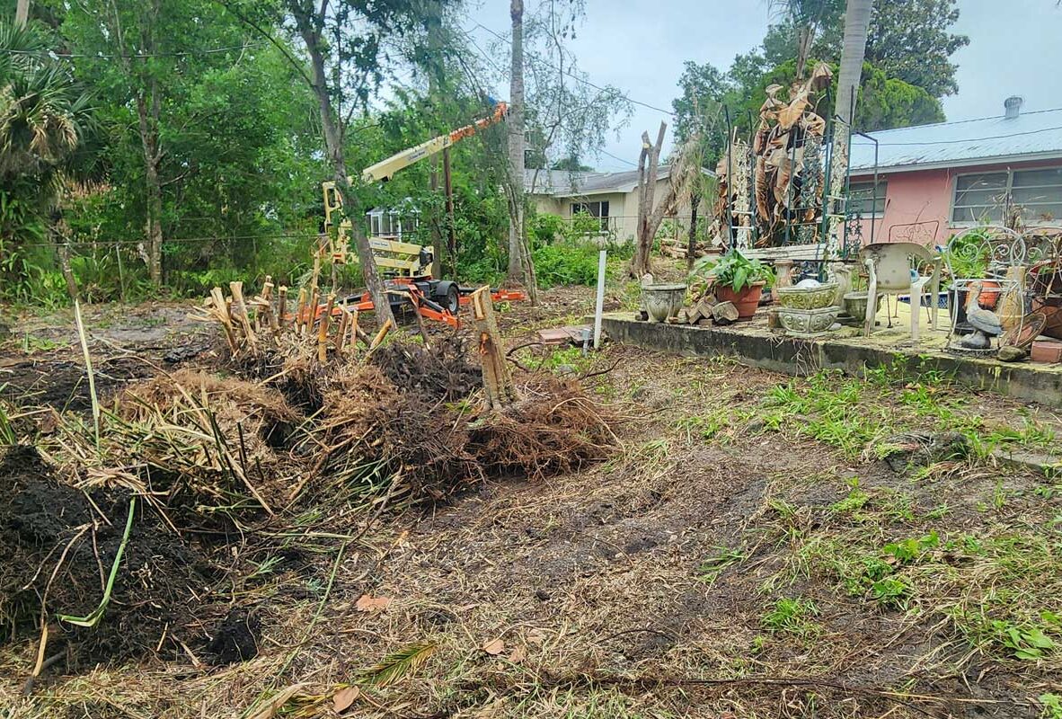 A backyard with uprooted vegetation and exposed soil; a tractor is parked near a pile of debris, and garden furniture sits by potted plants next to a house.